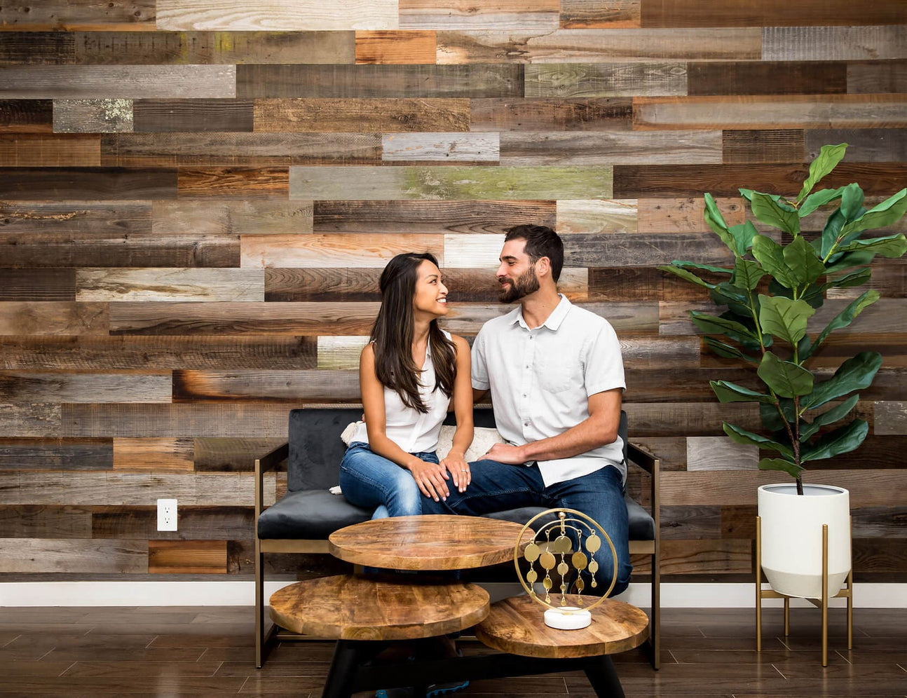 A couple sits on a modern bench against a reclaimed wood accent wall. The warm, earthy tones of the wood create a cozy backdrop. The room features a potted plant and a set of round wooden nesting tables.