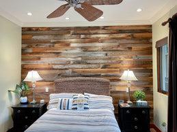 A cozy bedroom features a striking accent wall made of reclaimed wood planks in varying shades of brown. The room is decorated with a wicker headboard, black nightstands, and a ceiling fan with woven blades, creating a warm and rustic atmosphere.